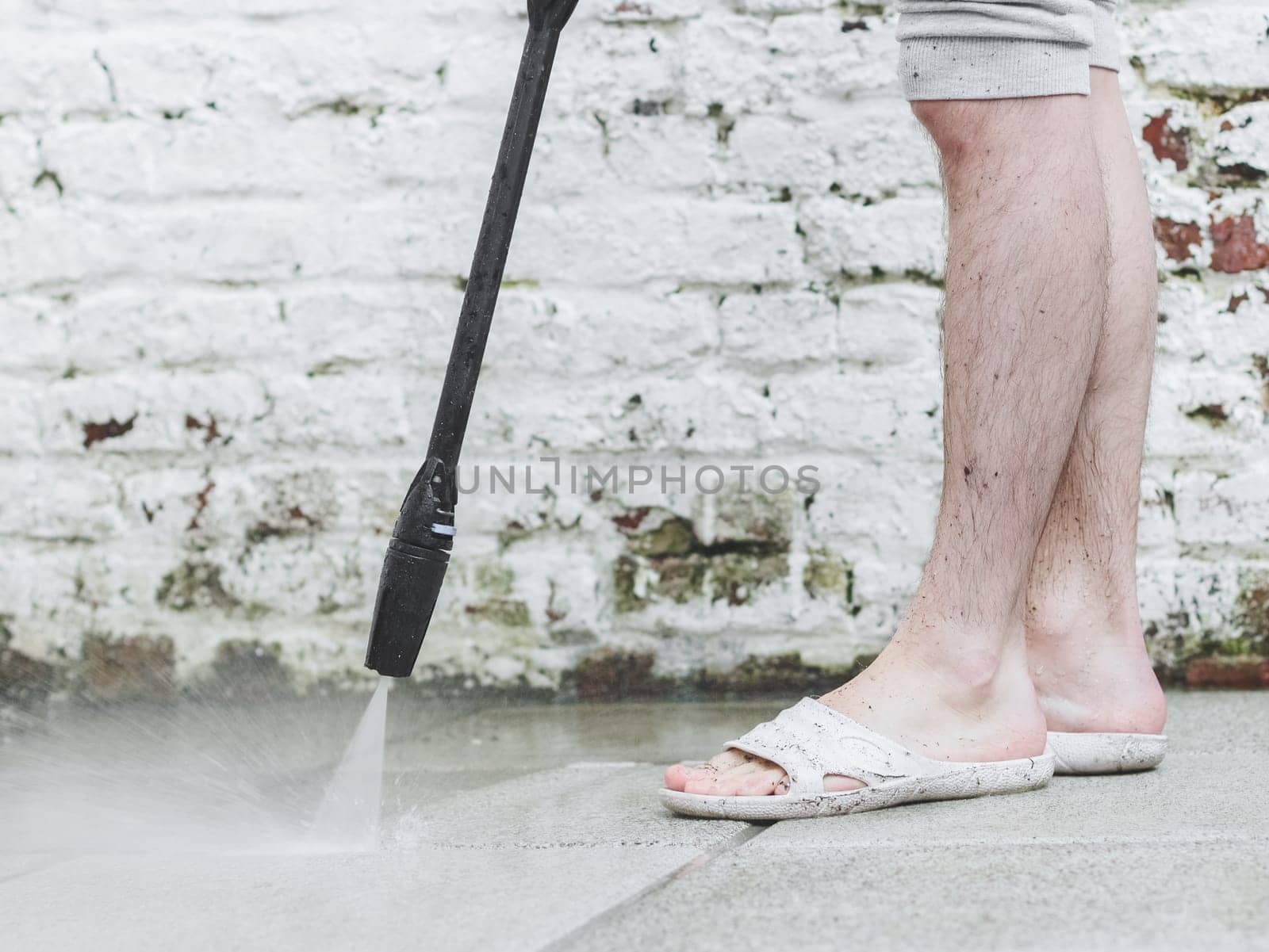 A caucasian man with hairy legs in white slippers washes the stone tiles of the terrace near the old painted brick wall in the backyard of his house under the pressure of water from the kercher, close-up. The concept of washing sun loungers, house cleaning services.