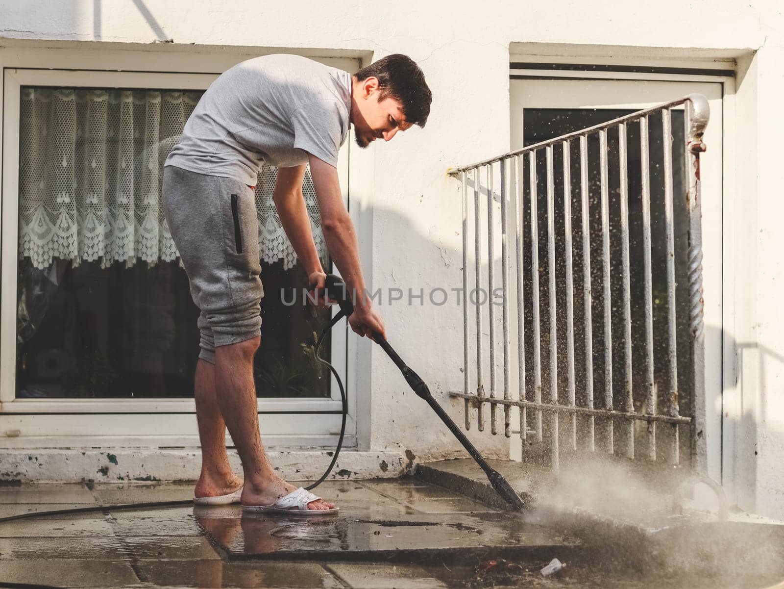 A caucasian young guy in gray sweatpants and a T-shirt washes with a karcher under the pressure of water with a frozen splash of stone tiles in the backyard of his house, side view close-up with depth of field.The concept of washing the yard,holiday preparation,at home,cleaning services,renovation.