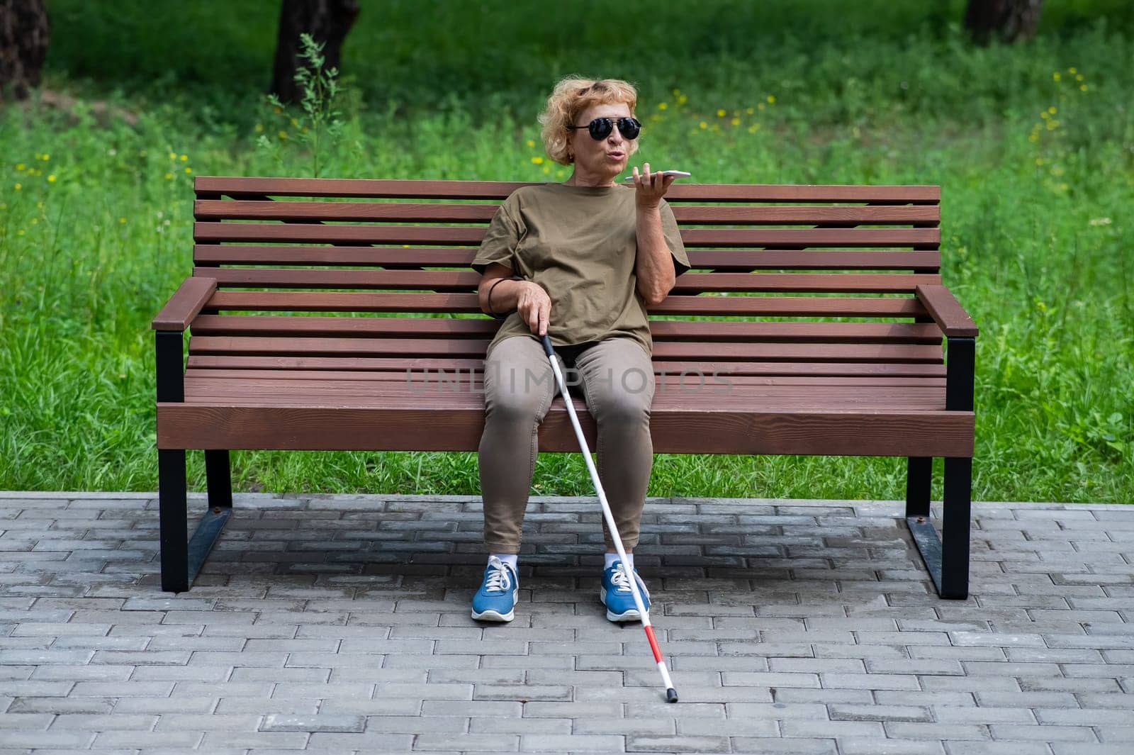 An elderly blind woman sits on a bench in the park and talks on a smartphone