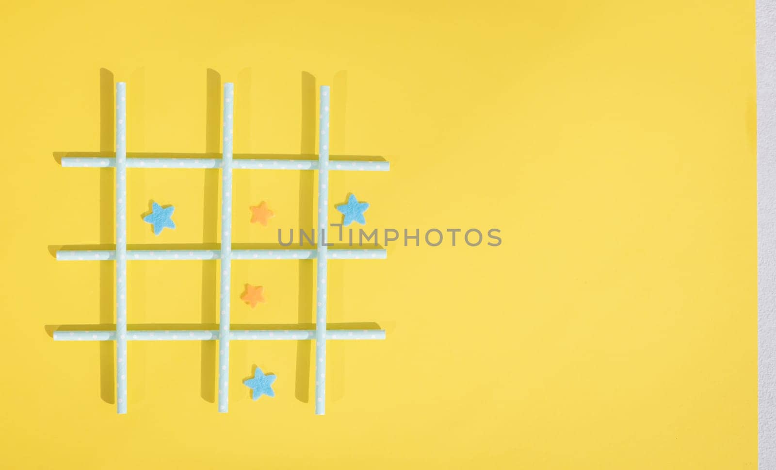 Blue straws with white polka dots lie in a lattice, and inside the squares, felt stars lie on the left on a yellow background with copy space on the right, flat lay, close-up. Concept summer board games.