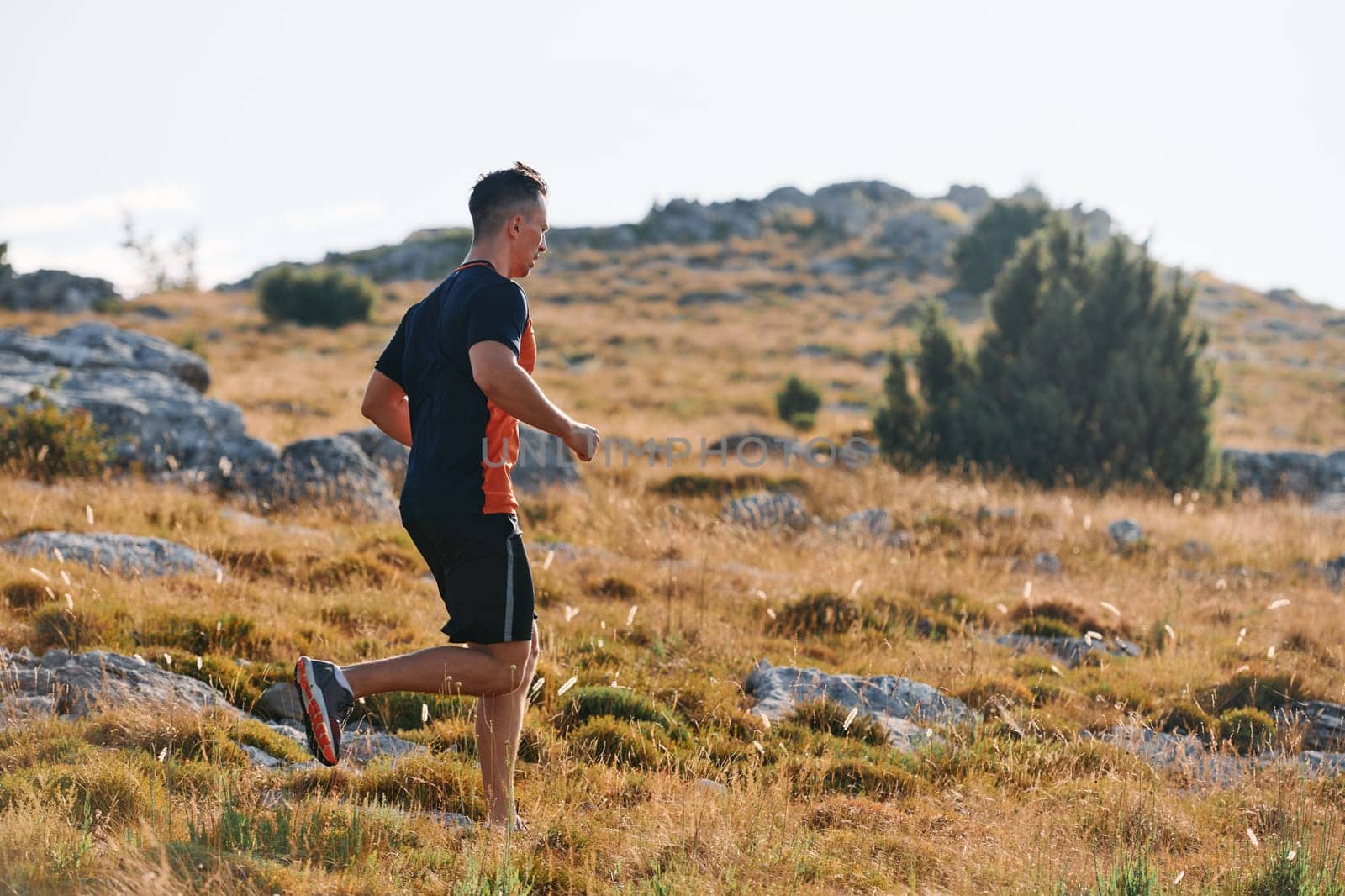 A muscular male athlete runs along a rugged mountain path at sunrise, surrounded by breathtaking rocky landscapes and natural beauty.