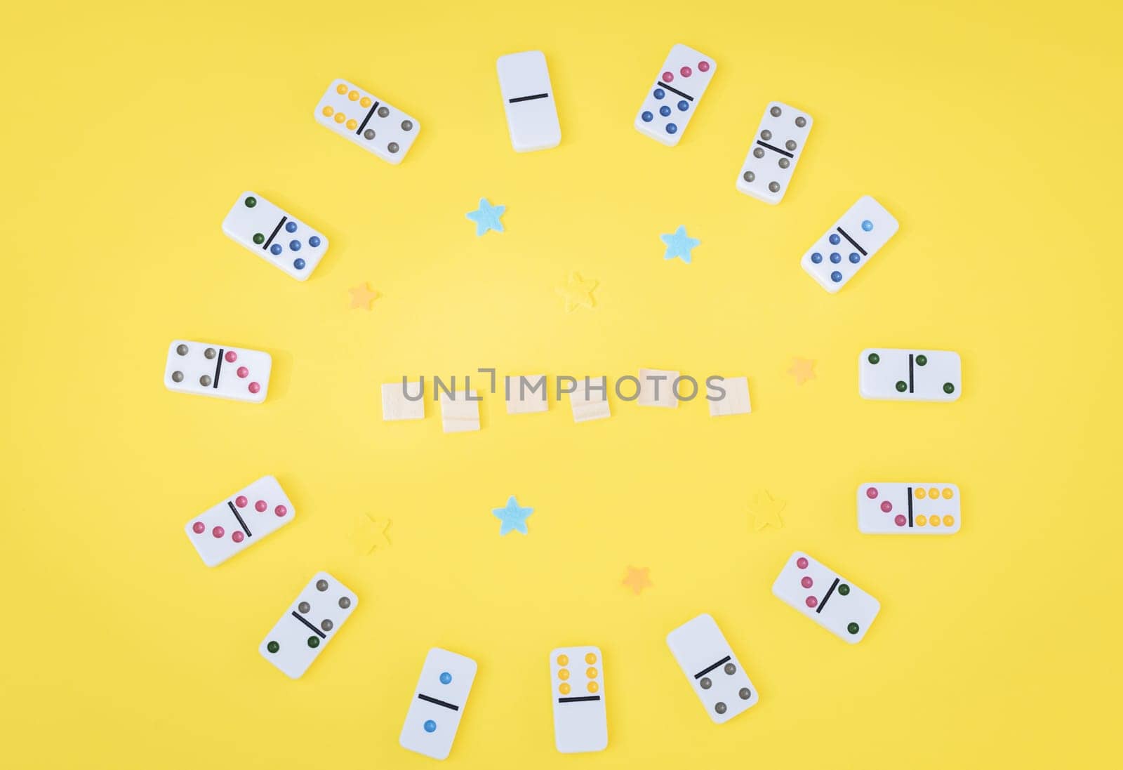 White dominoes with colored dots and empty wooden cubes are arranged in a circle on a yellow background with copy space in the center, closeup flat lay. Summer board game concept.