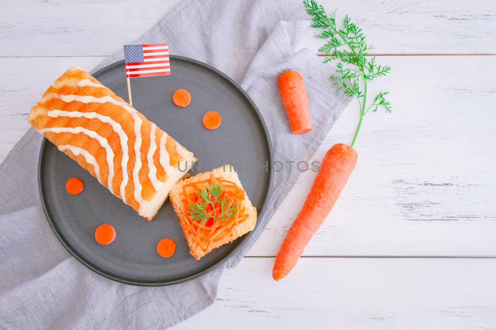 A delicious cake with sugar icing, fresh carrots, American flag paper and a cut piece in a plate lies on a wooden table on the left with space for text on the right, top view, close-up. Carrot cake national day concept.