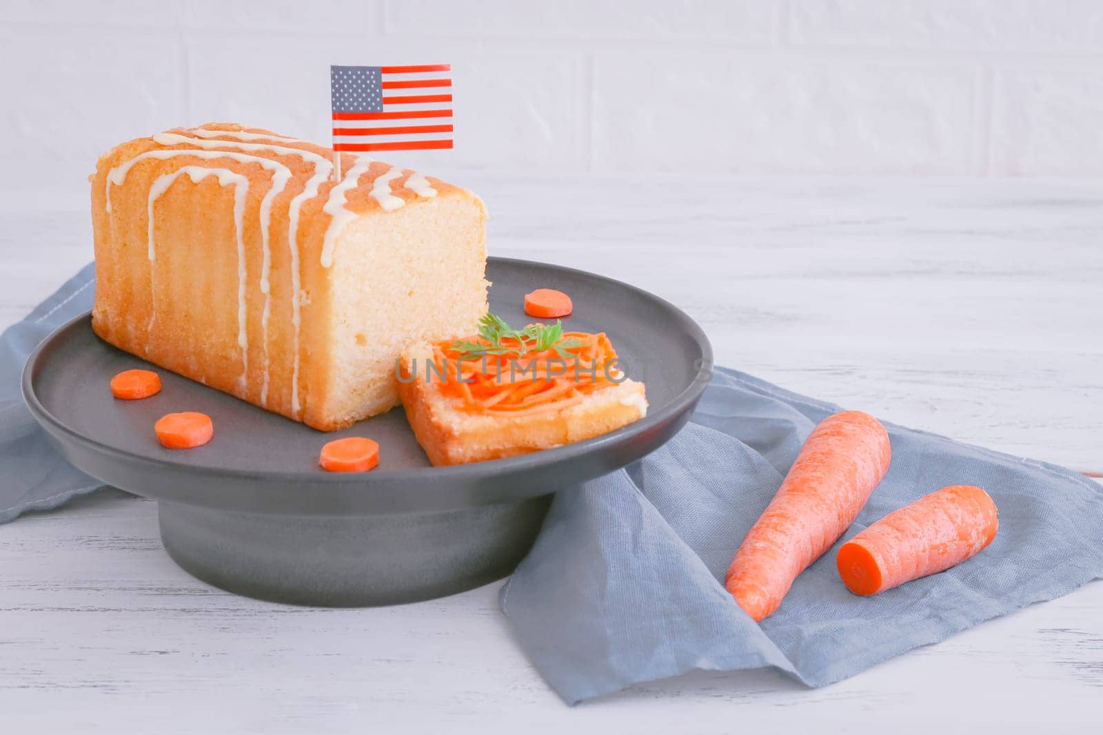 Carrot cake with cut off piece, american flag paper, on gray porcelain cake stand with gray kitchen napkin on white shabby wooden table, closeup side view.