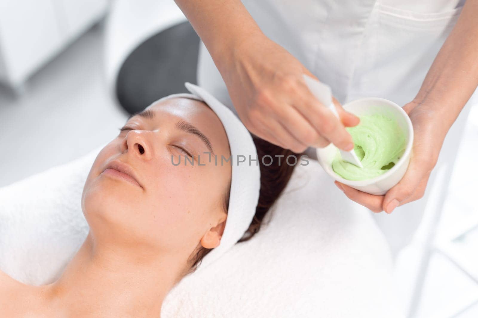 Beautician mixing creamy substance in bowl. Cosmetologist preparing alginate cream for relaxed woman lying in beauty salon. Spa and beauty treatment concept.