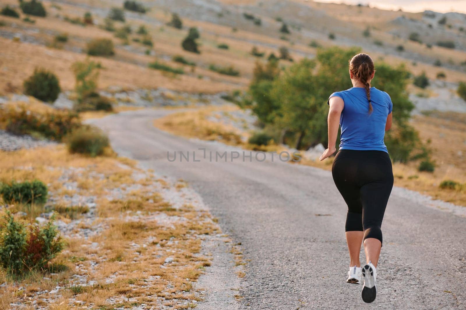 A determined female athlete runs through a forest trail at sunrise, surrounded by breathtaking natural beauty and vibrant greenery.