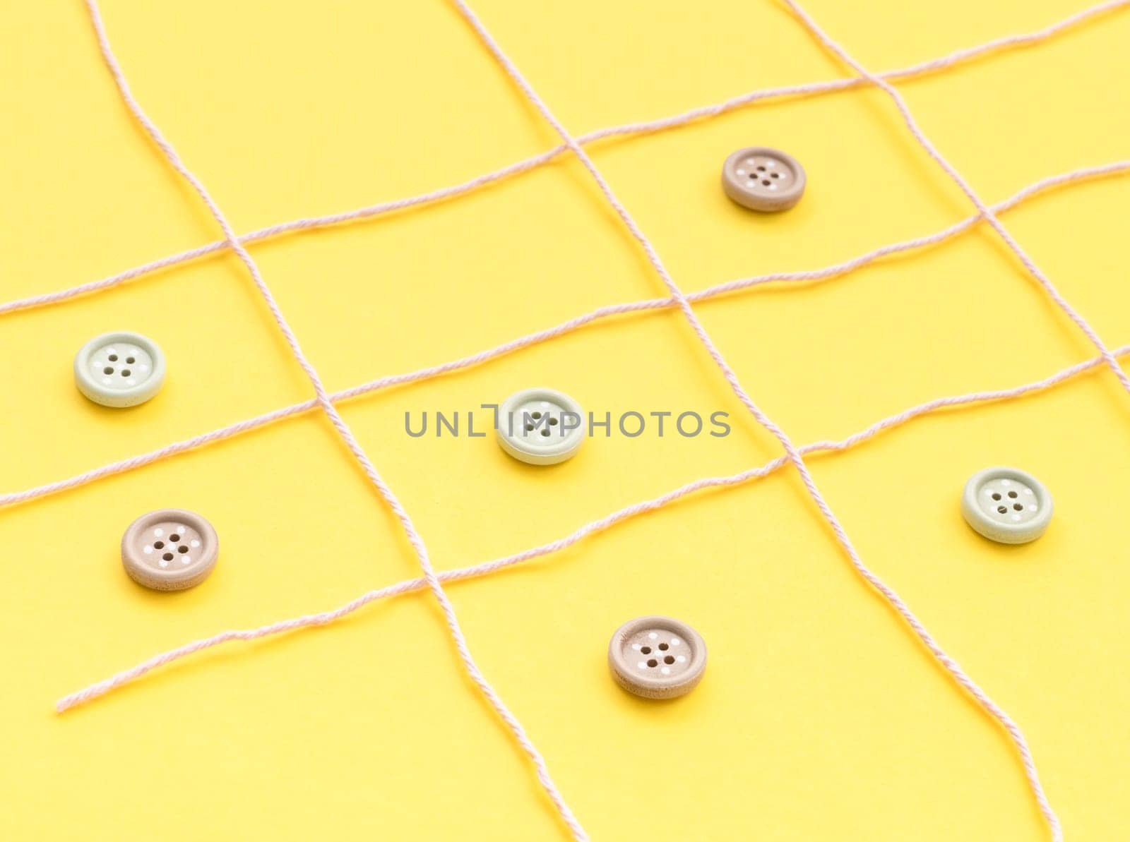 Pale lilac woolen threads lie in a lattice, and inside the squares there are buttons on a yellow background, side view, close-up. Concept summer board games.