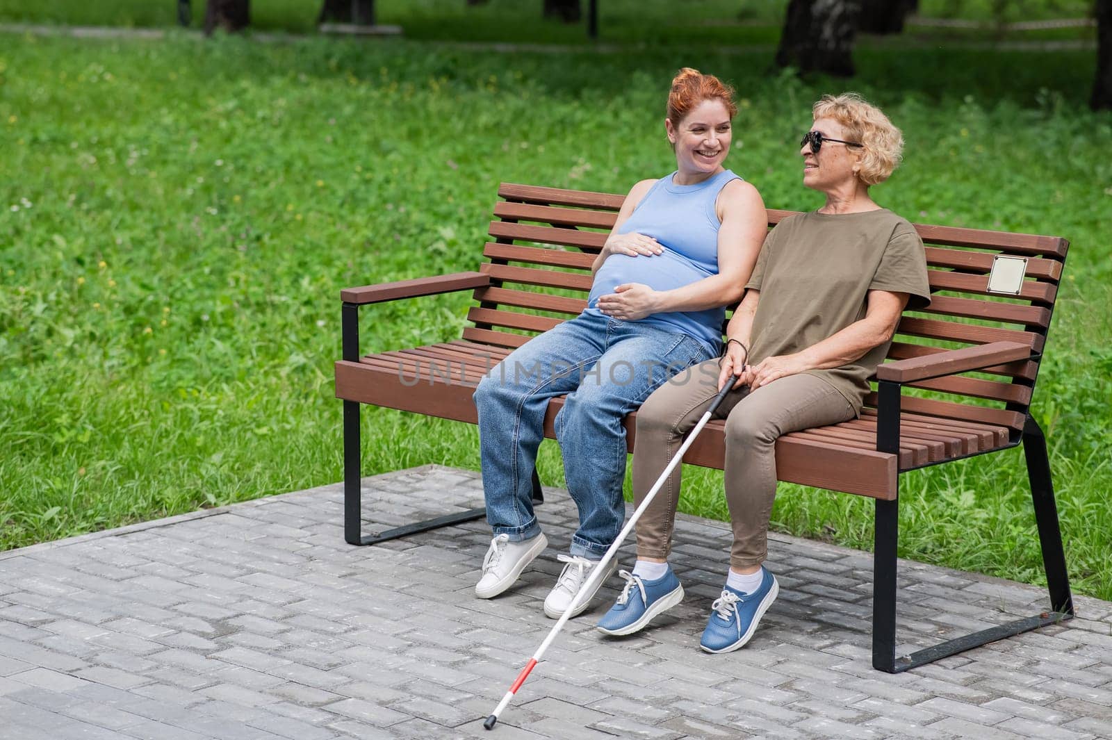 An elderly blind woman and her pregnant daughter are sitting on a bench in the park