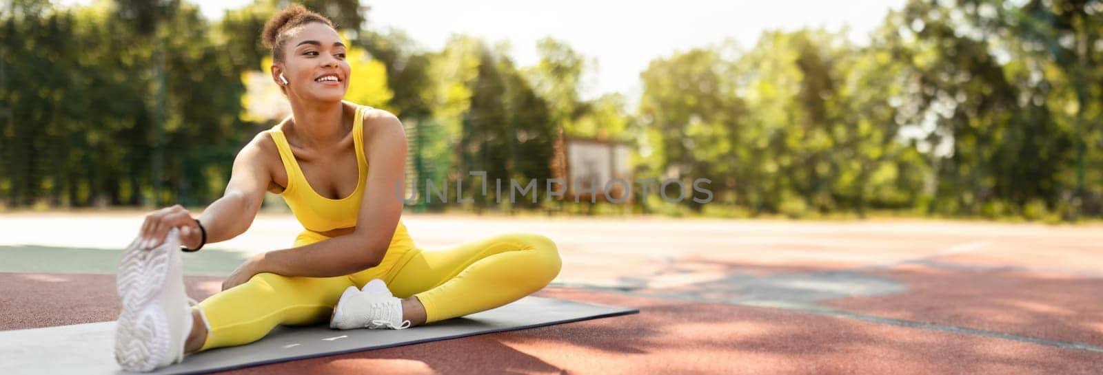 Sporty Lifestyle Concept. Smiling African American lady in yellow top, leggings and white sneakers stretching back, arms and leg muscles sitting on yoga mat outside, touching feet, copy space
