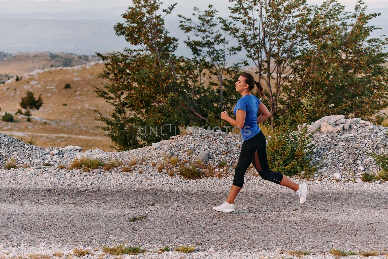 A determined female athlete runs through a forest trail at sunrise, surrounded by breathtaking natural beauty and vibrant greenery.