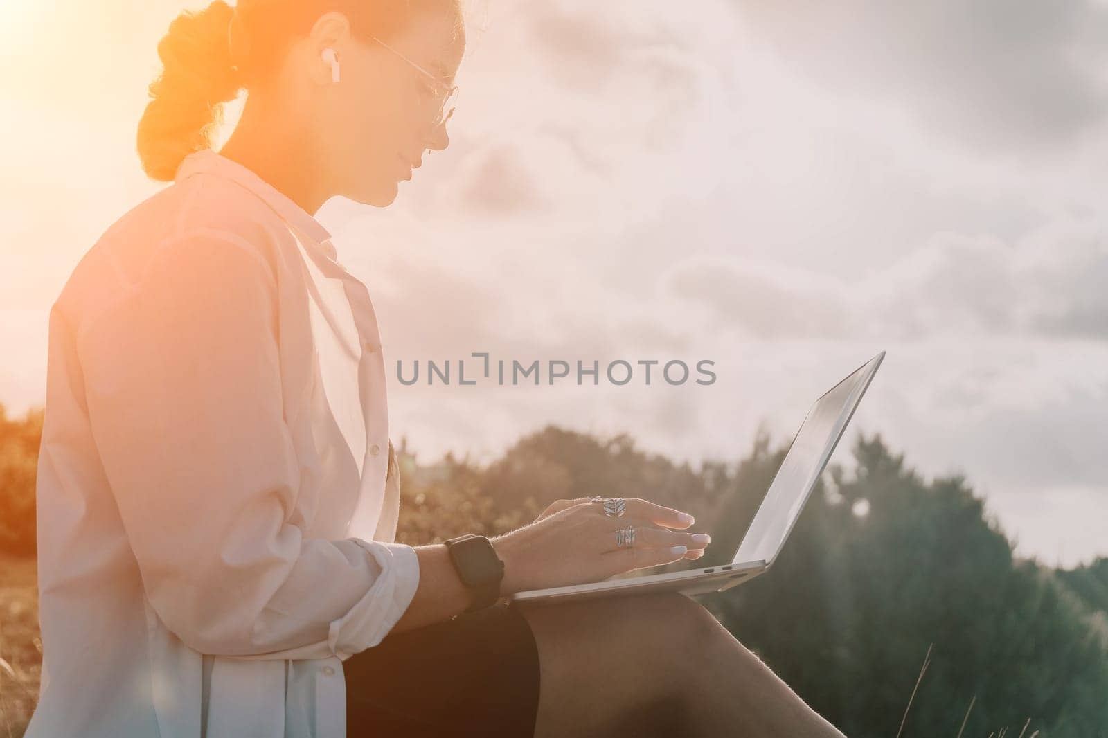 Digital nomad, woman in the hat, a business woman with a laptop sits on the rocks by the sea during sunset, makes a business transaction online from a distance. Freelance, remote work on vacation.