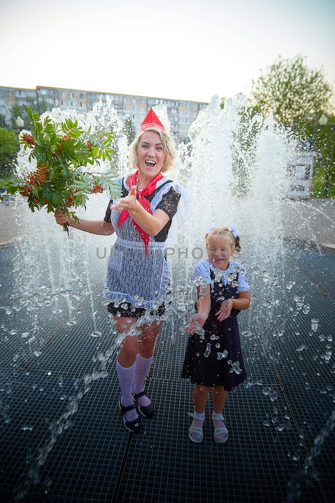 Young and adult schoolgirl on September with flowers having fun near water of fontain. Generations of schoolchildren, pioneer of USSR and October girl in modern uniform of Russia. Mother and daughter by keleny