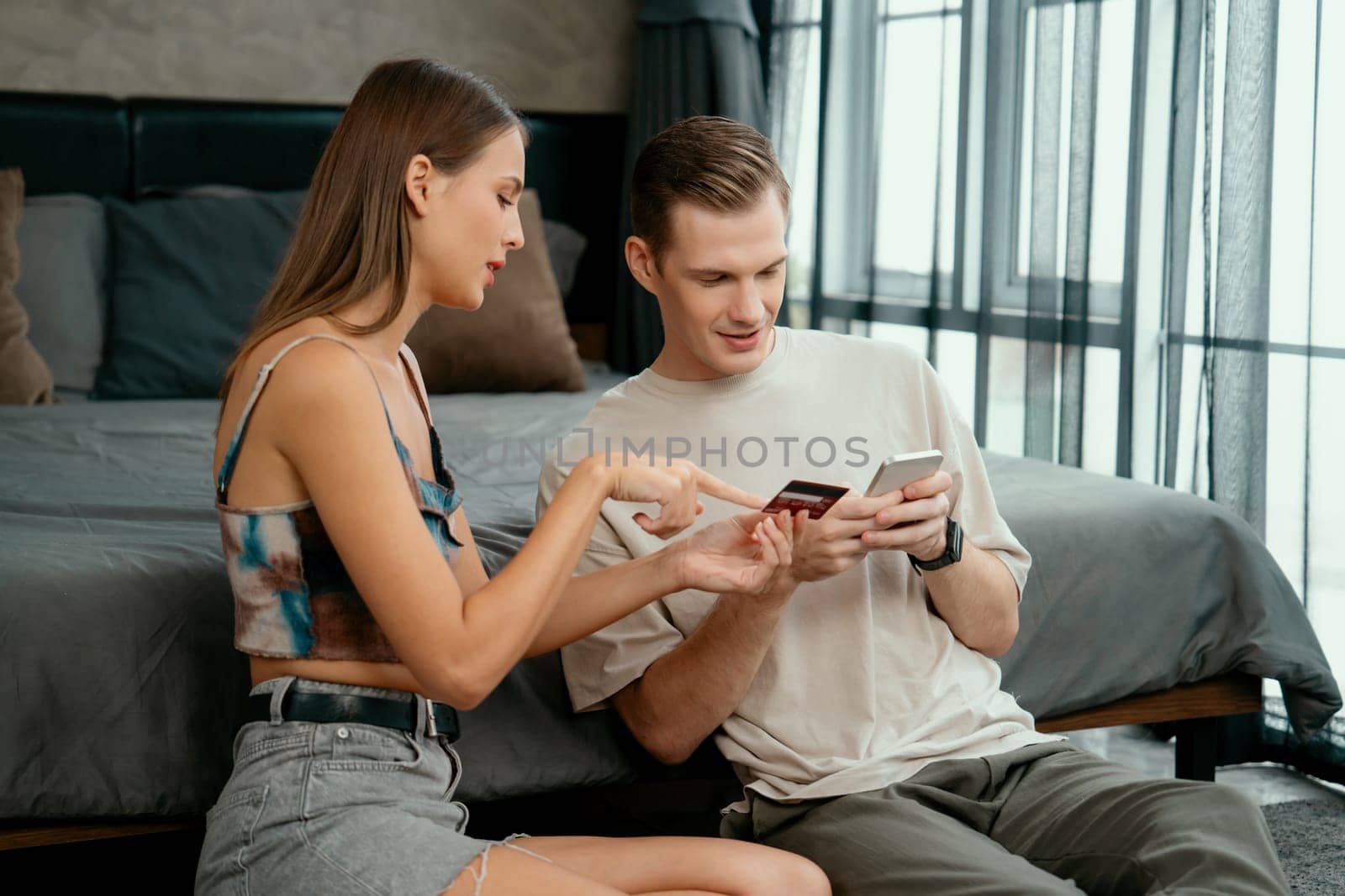 Young couple sit in the home bedroom using online payment app and digital wallet on smartphone to pay with credit card. E commerce shopping and modern purchasing via mobile internet. Unveiling