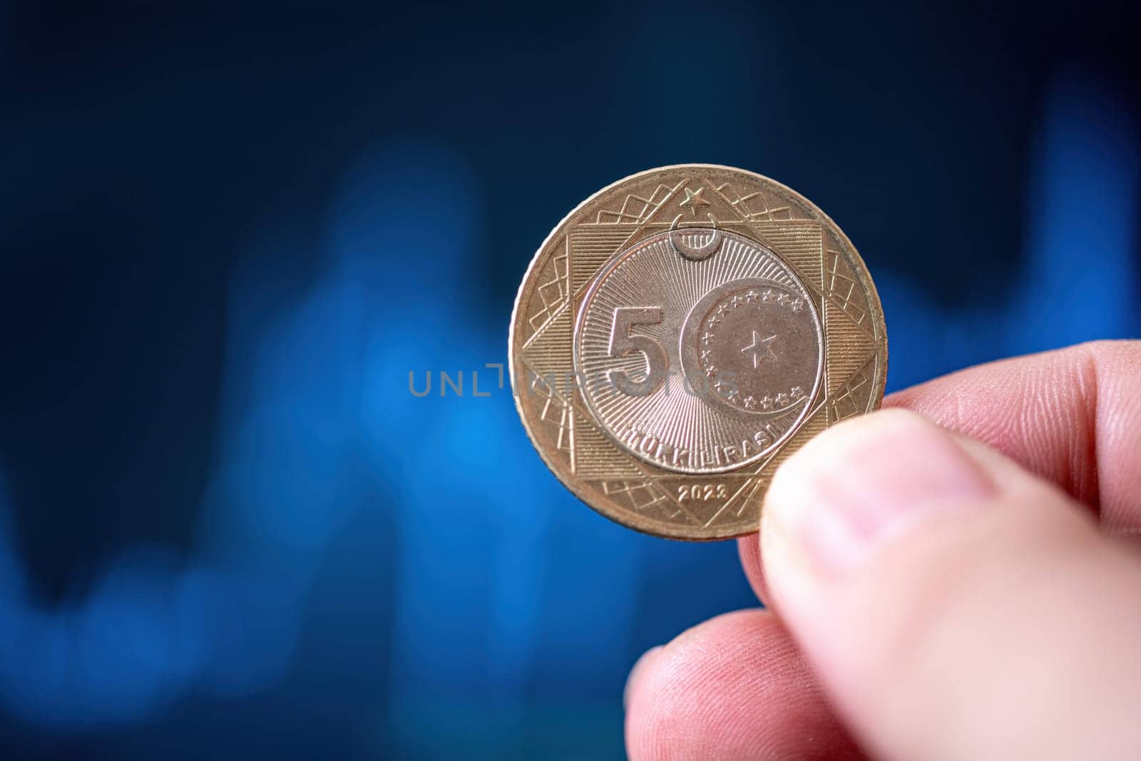 Hand holding a 5 Turkish Lira coin in front of a screen with financial chart curves by Sonat