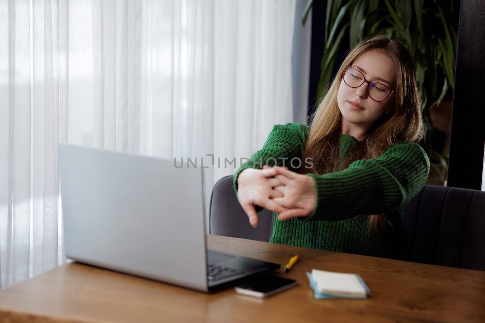 A woman is sitting at a desk in front of a laptop computer, stretching her arms. Keywords Table, Computer, Laptop, Wood, Gesture, Personal computer, Flooring, Hardwood, Wood stain, Thumb