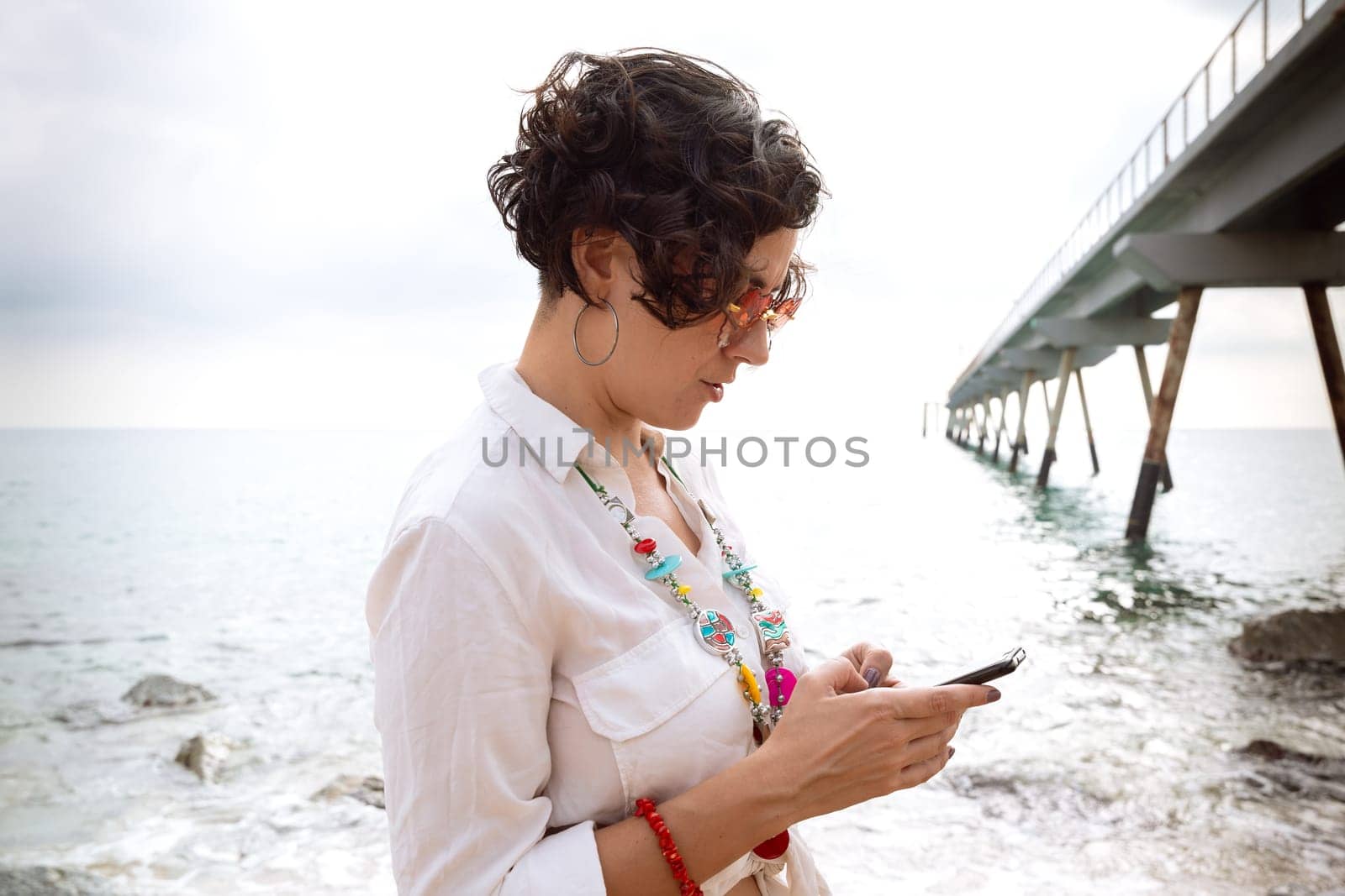 caucasian woman wearing sunglasses on vacation by the sea laughing using a social media app with her mobile phone, holding a caffee.