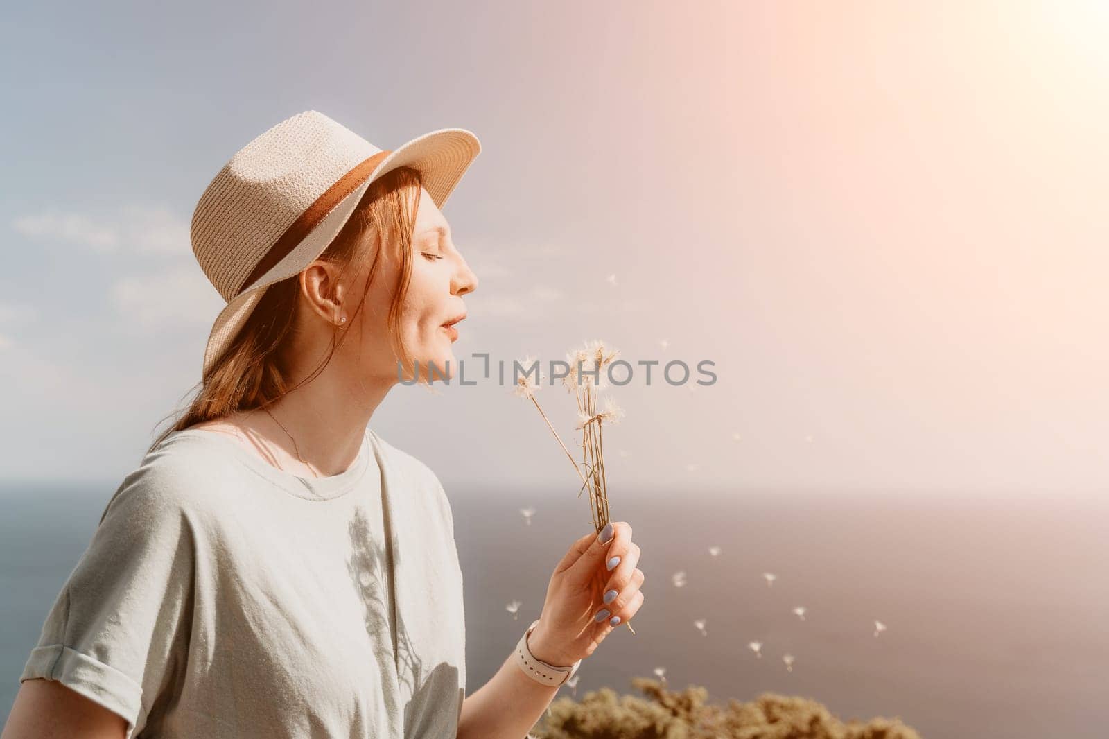 Woman travel sea. Happy tourist in hat standing on a beach and holding a bouquet of dandelion flowers. She is blowing the petals into the air, creating a beautiful and serene scene. by panophotograph