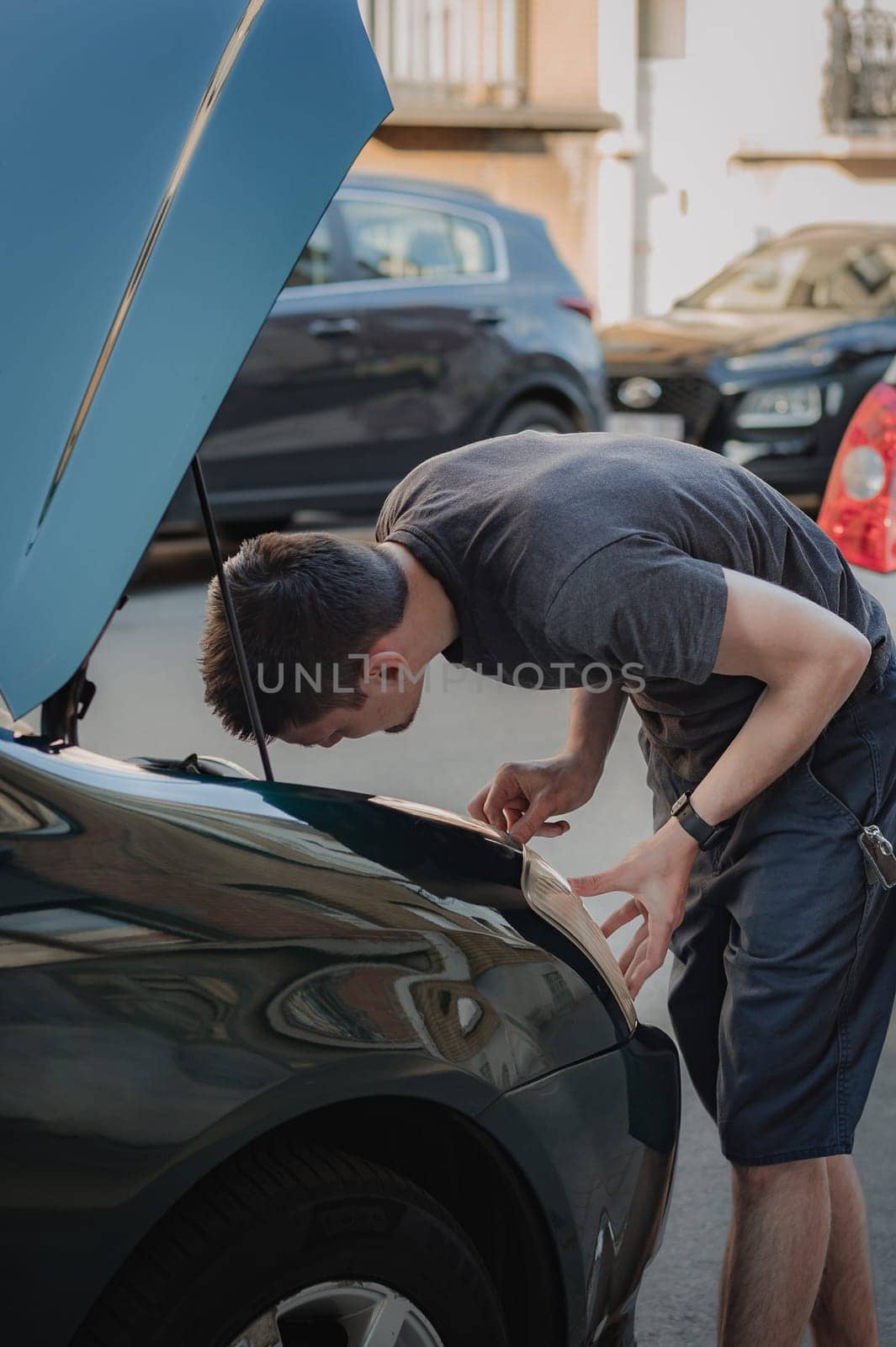 One young caucasian guy bending over checks the front right headlight of his car, looking into the open hood, standing on a city street on a summer afternoon in the evening, close-up side view. The concept of replacing light bulbs, car repair.