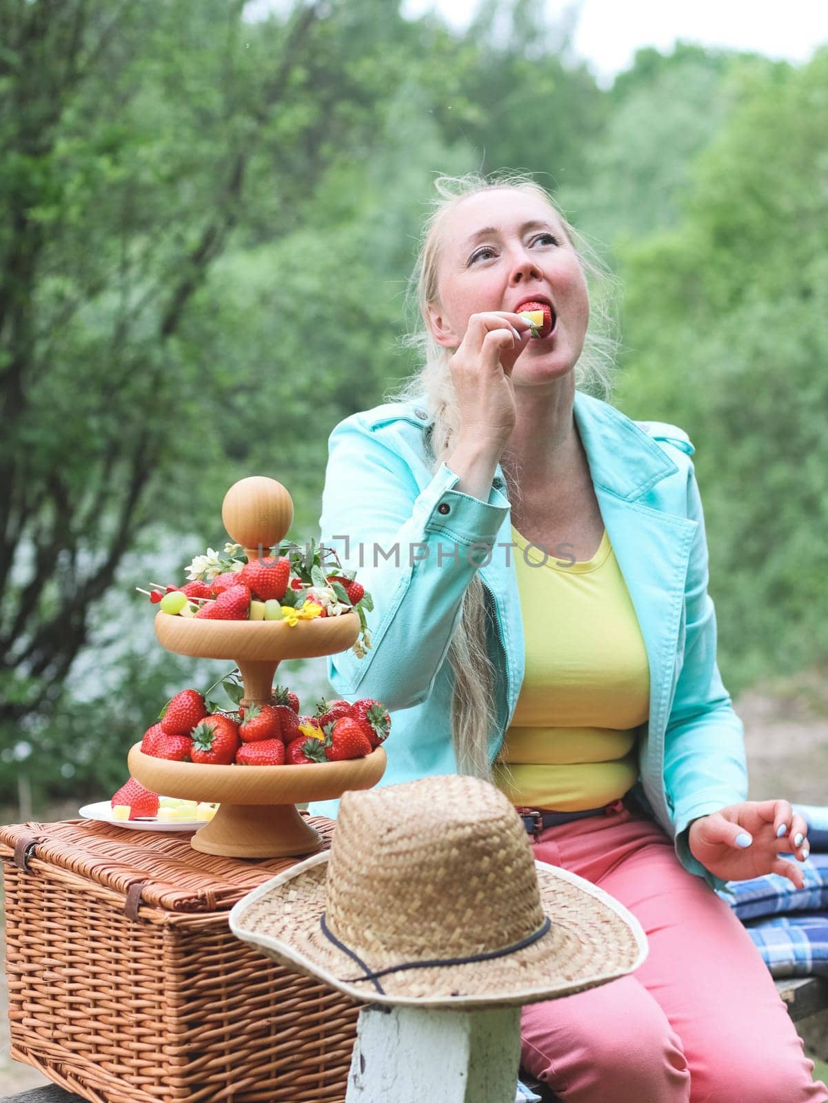 Portrait of a beautiful Caucasian cheerful middle-aged blonde woman sitting on a bench with a wicker basket, a wooden two-tier plate and eating a fruit barbecue on a skewer while sitting in a park on a spring day, close-up side view. Outdoor picnic concept, healthy eating and lifestyle.