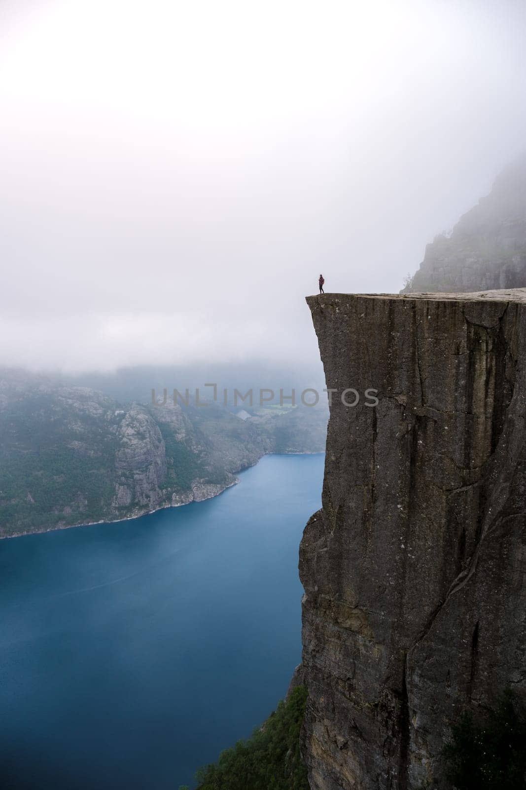 Preikestolen, Norway, A lone figure stands on the edge of a dramatic cliff overlooking a fjord in Norway. The misty landscape and the vast expanse of water create a sense of awe and isolation.