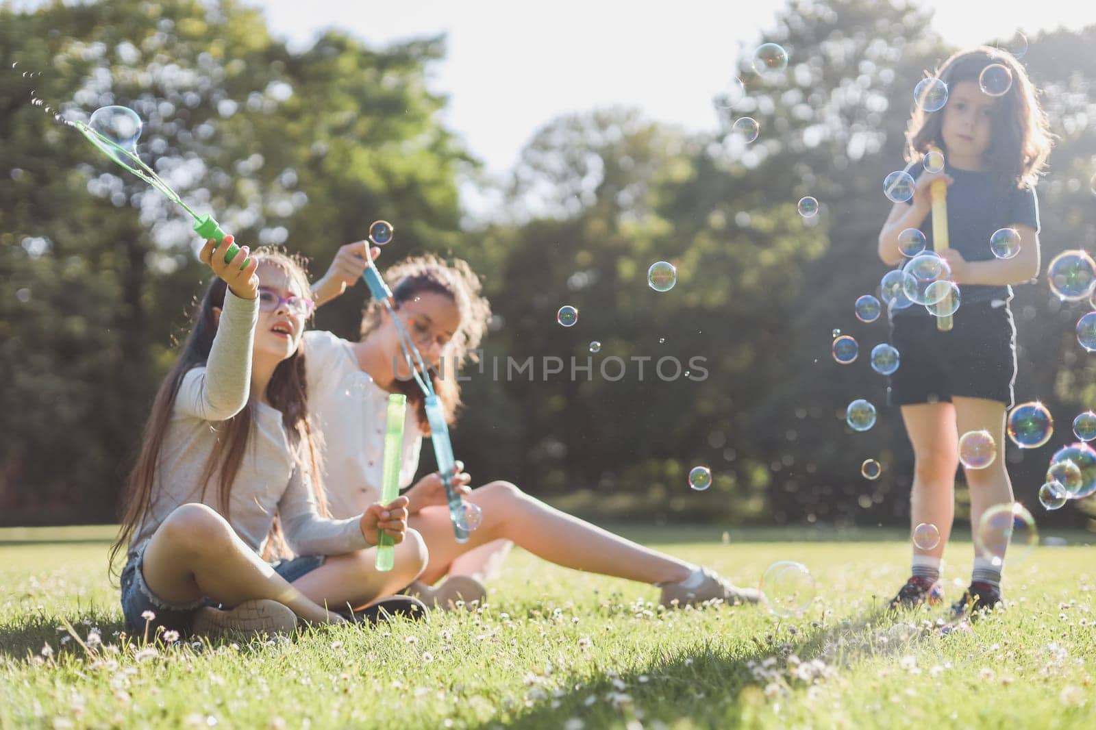 Three beautiful caucasian girls sisters are sitting on a flower lawn in a public park and blowing soap bubbles in the backlight with a blurred background, close-up side view. PARKS REC concept, happy childhood, children picnic,holidays,children recreation,outdoors.