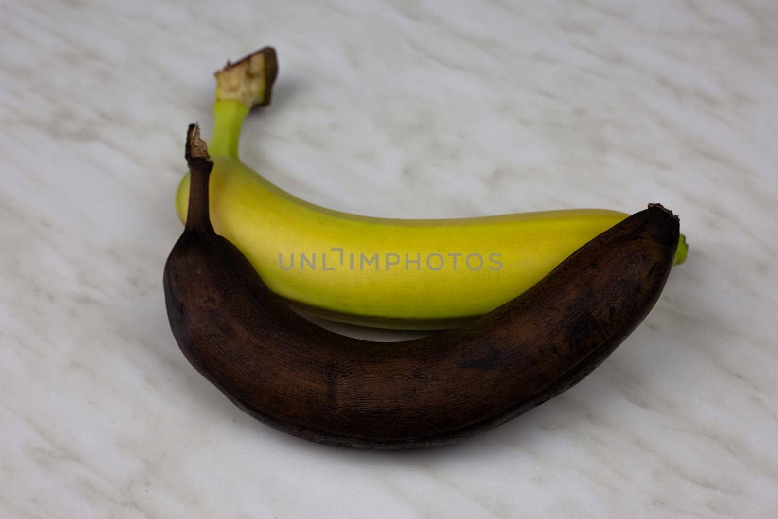 Spoiled rotten banana lies on fresh banana, expired banana fruit in peel, black banana on white table