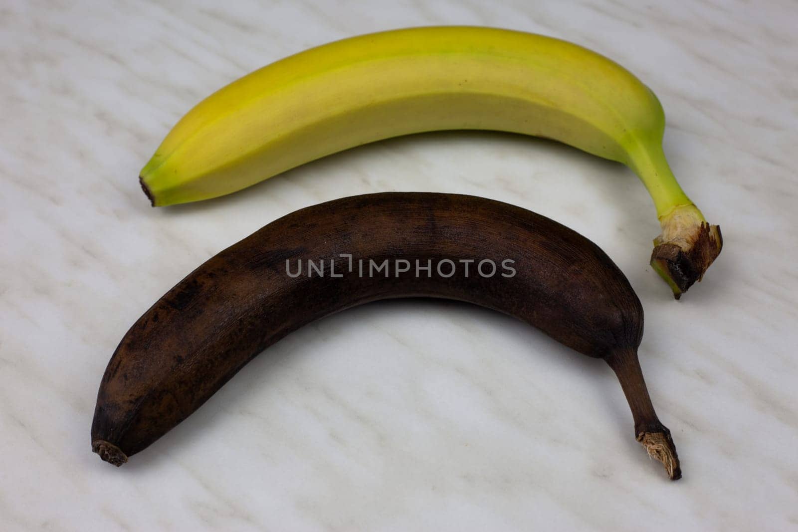 Rotten banana in a peel lies on white table with fresh fruit by timurmalazoniia