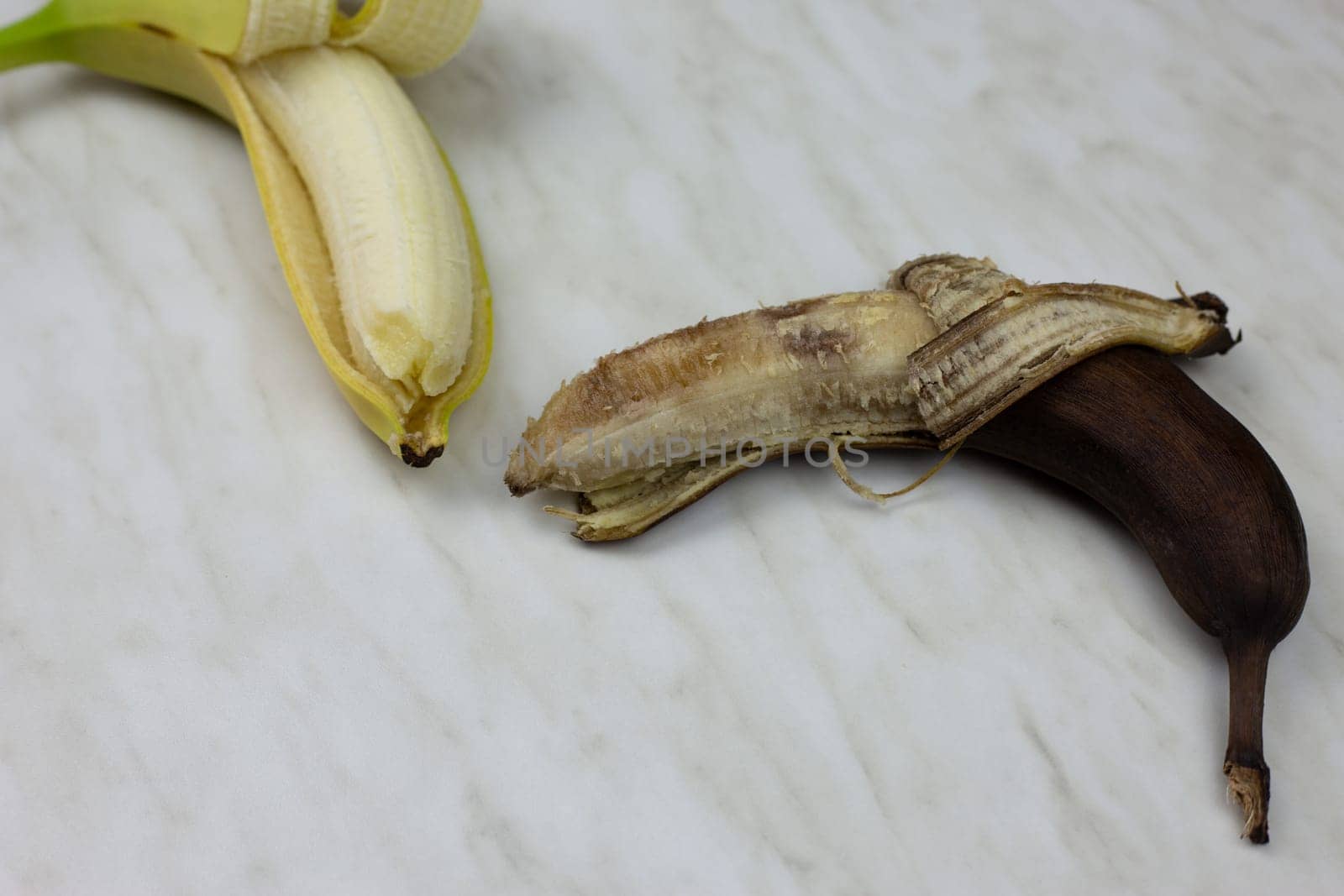 Rotten spoiled banana in open peel next to fresh banana fruit on white table, phase of spoiled fruit