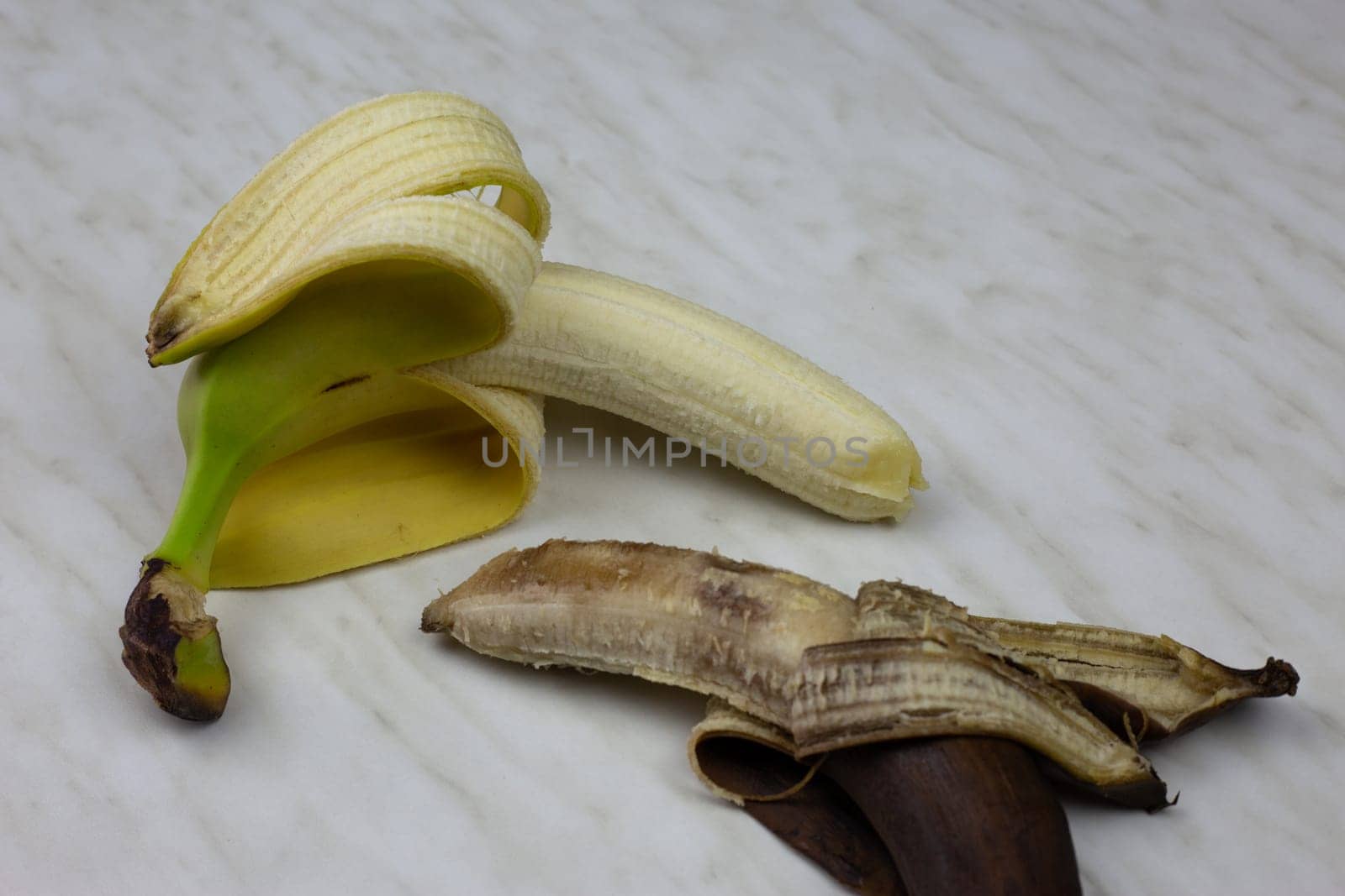 Juicy fresh banana fruit in open yellow peel next to rotten expired banana on white table, ugly brown banana not edible with fresh fruit