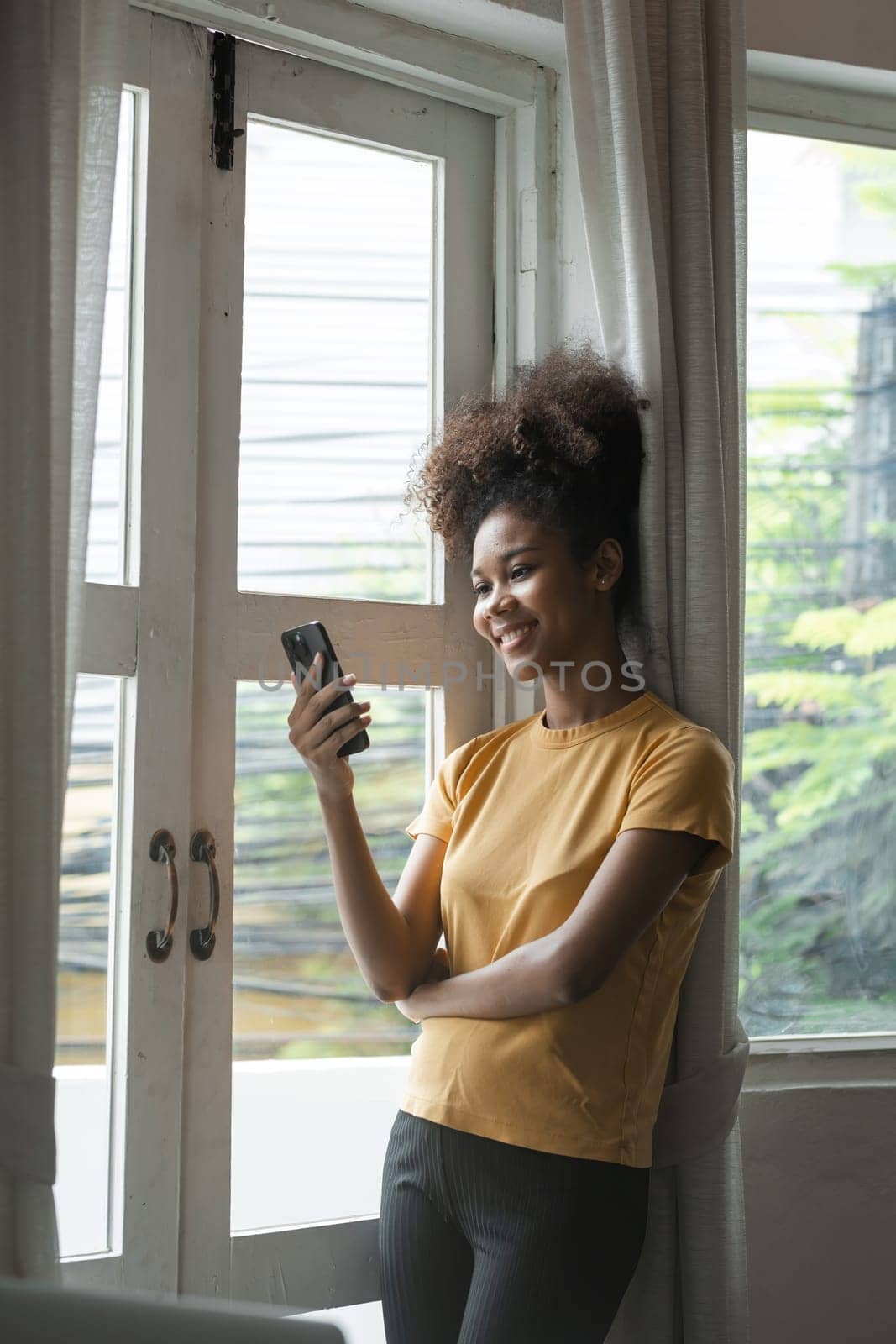 Young African Woman Smiling While Using Smartphone at Home by the Window, Casual Clothing, Natural Light, Modern Lifestyle, Relaxed and Happy, Indoor Setting, Daytime, Technology and Communication by wichayada