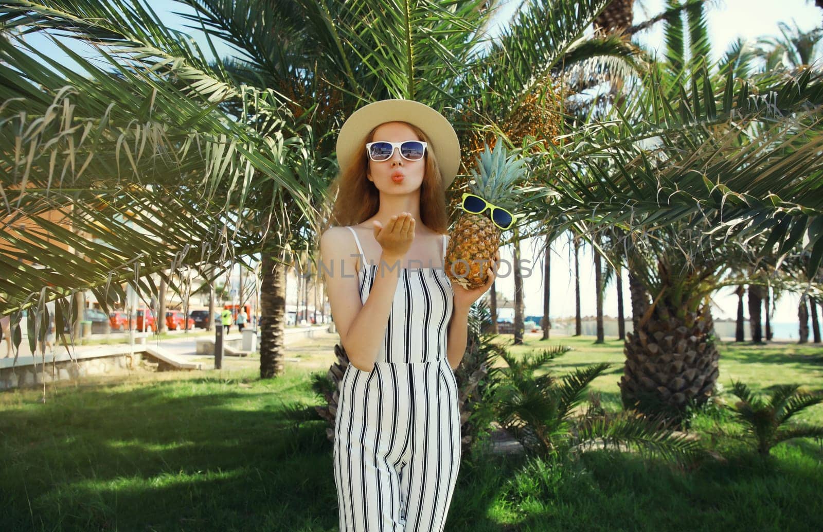 Summer fashion outfit of trendy young woman blowing a kiss holding pineapple fruits wearing hat, striped clothes, sunglasses posing in park with palm trees background