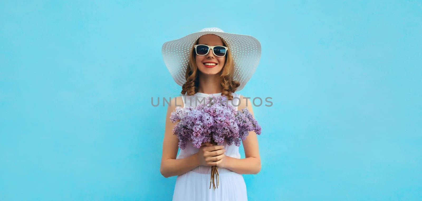 Summer portrait of beautiful lovely happy smiling woman with bouquet of purple lilac flowers in white straw hat on blue background