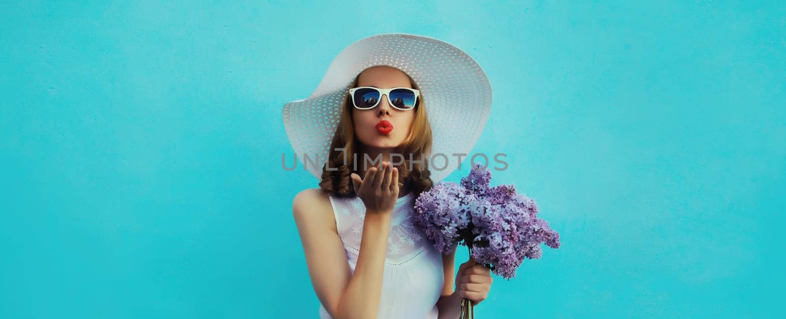 Summer portrait of beautiful lovely young woman blowing kiss with bouquet of purple lilac flowers in white straw hat on blue background