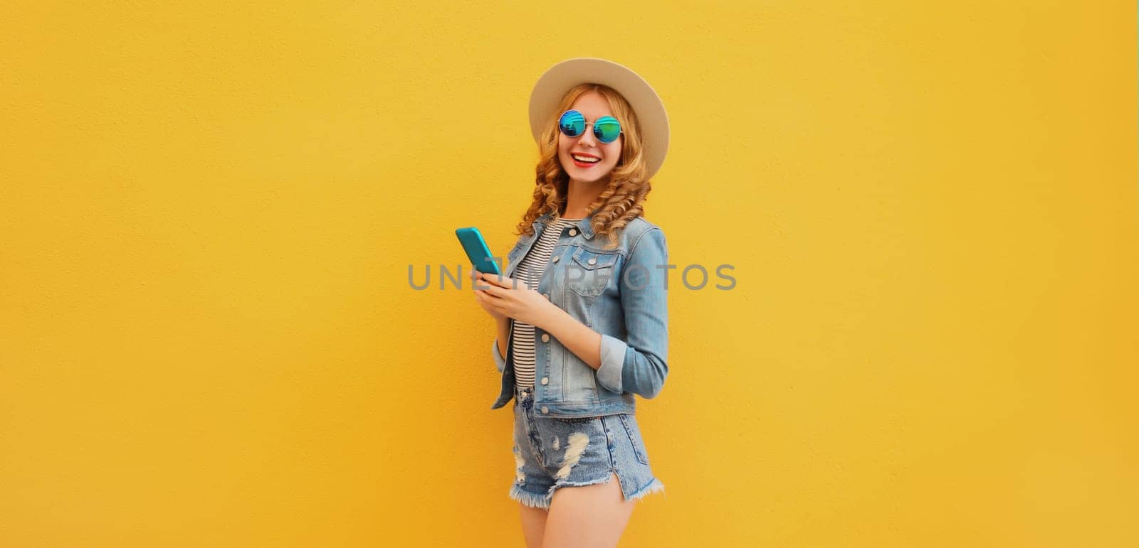 Summer modern happy young woman holding a smartphone in straw tourist hat, denim clothing posing on yellow wall background