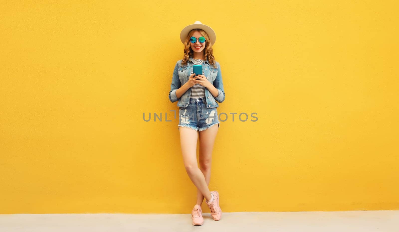 Summer trendy happy young woman with smartphone in straw tourist hat posing on yellow background by Rohappy