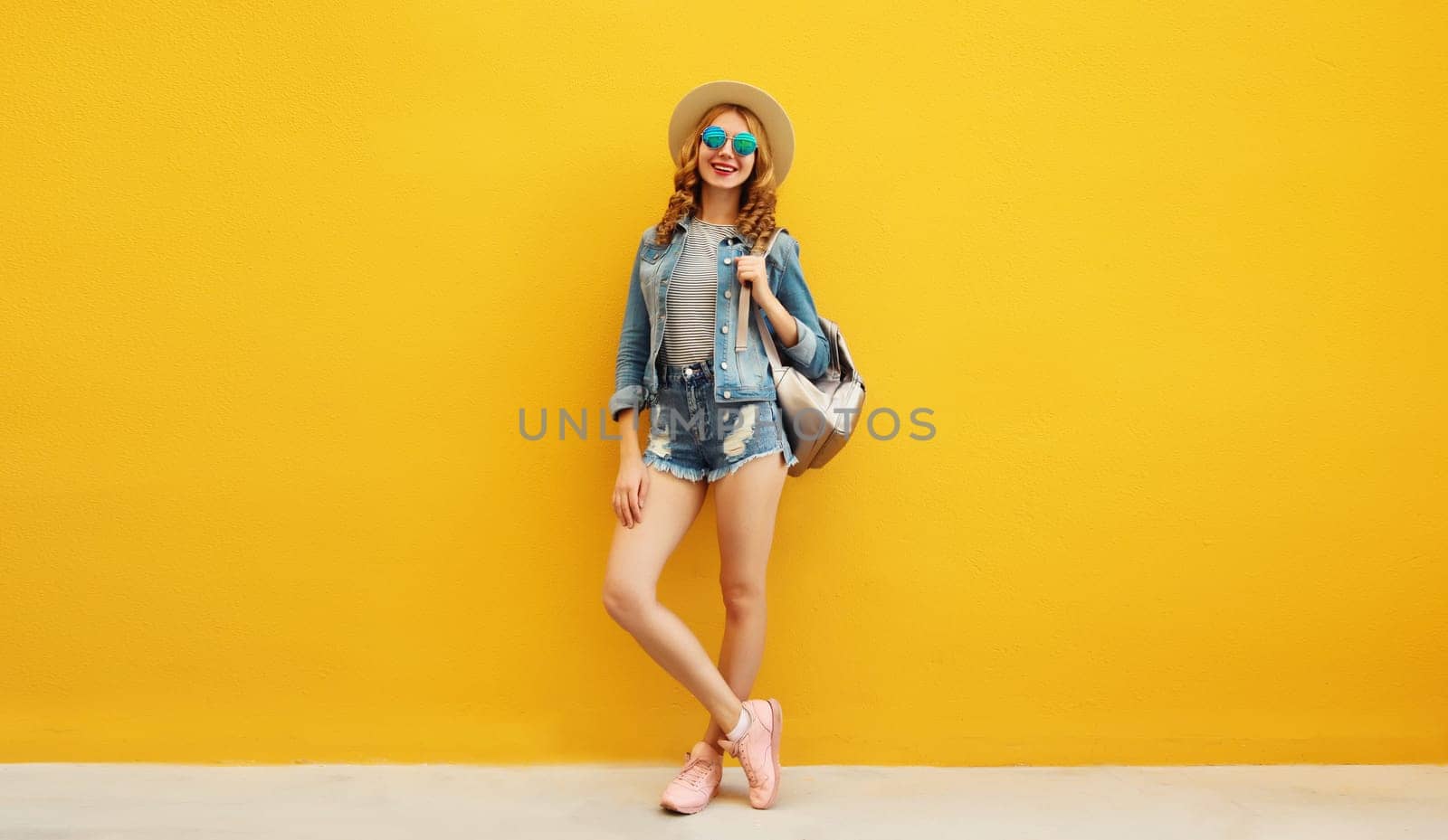 Beautiful young woman full length standing in summer straw hat posing with backpack, denim clothing on vivid yellow background