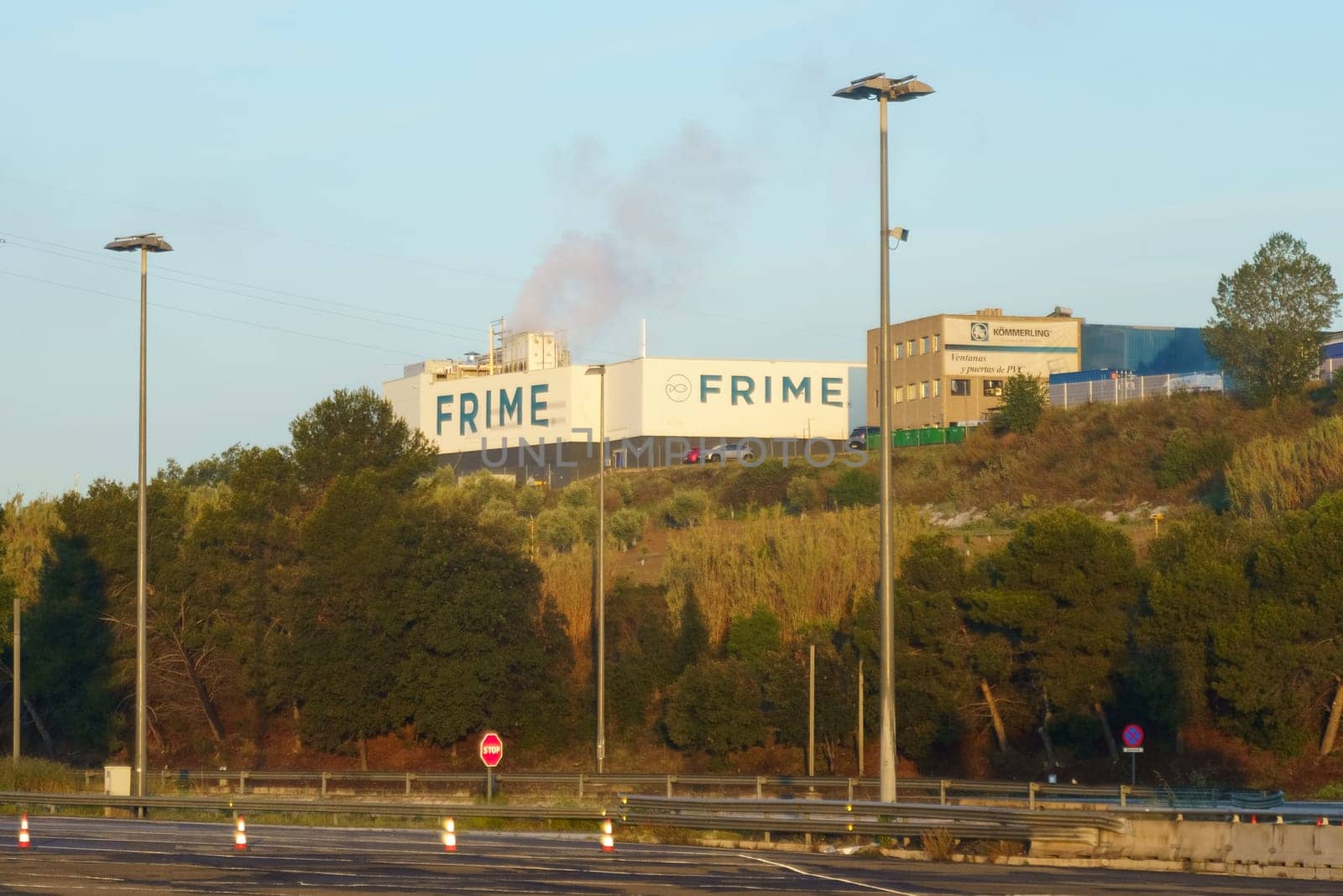 Barcelona, Spain - May 15, 2023: Smoke emerges from the FRIME factory behind a tree line as the sun sets, viewed from an empty highway.