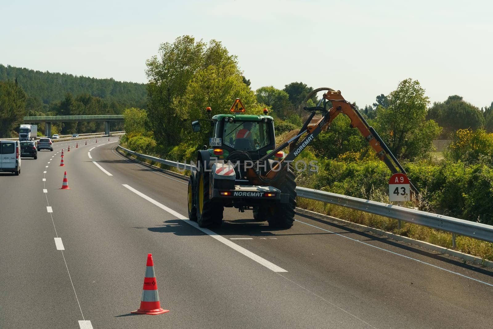 Salles-d'Aude, France - May 16, 2023: A maintenance worker operates an excavator on the side of a highway, with traffic cones set up for safety during the daytime.