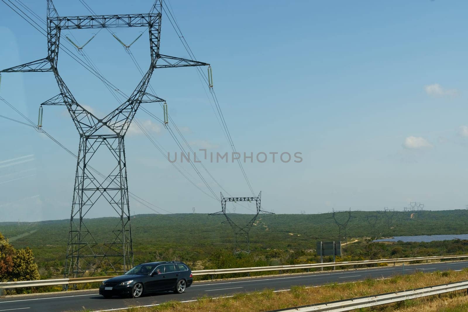 A black car drives on a highway, passing under a large power line tower in a rural landscape. The sky is blue and clear.