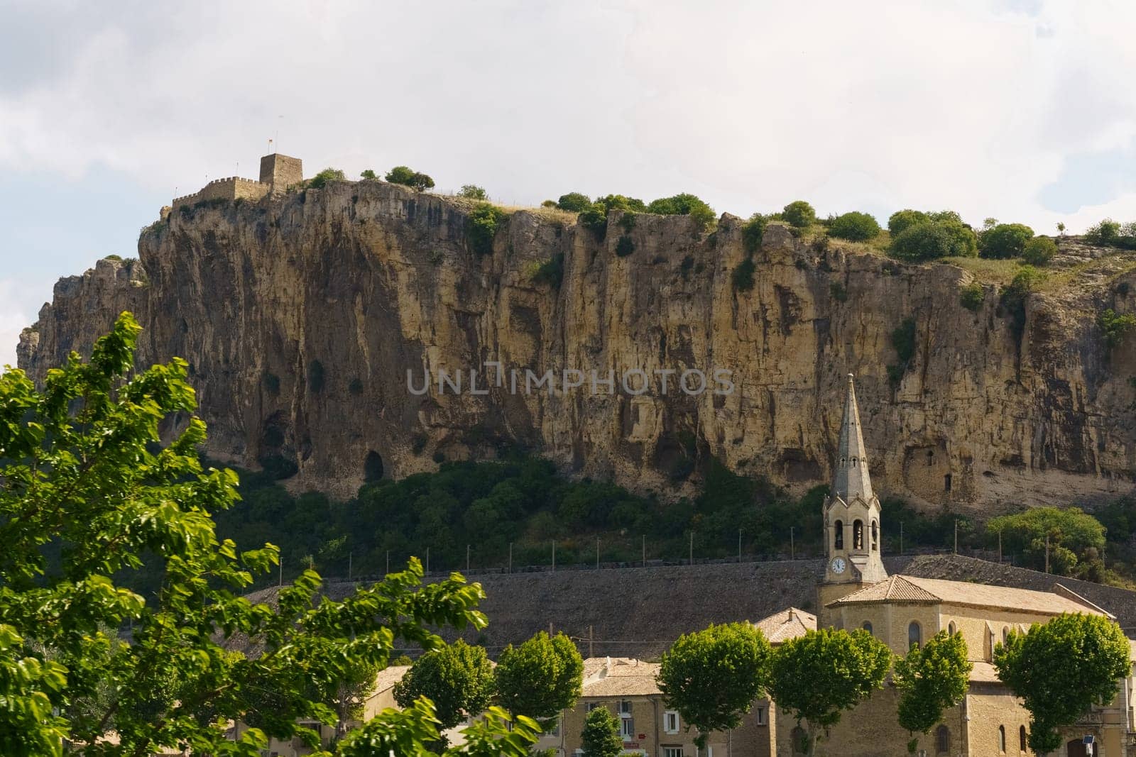 A church with a tall spire sits among buildings at the base of a large limestone cliff in a rural French town.