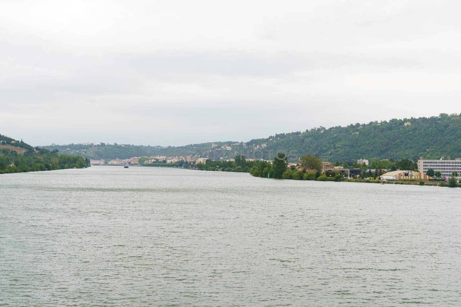 A wide shot of the Isere River flowing through the city of Grenoble, France, with a view of the surrounding mountains in the background on a cloudy day.