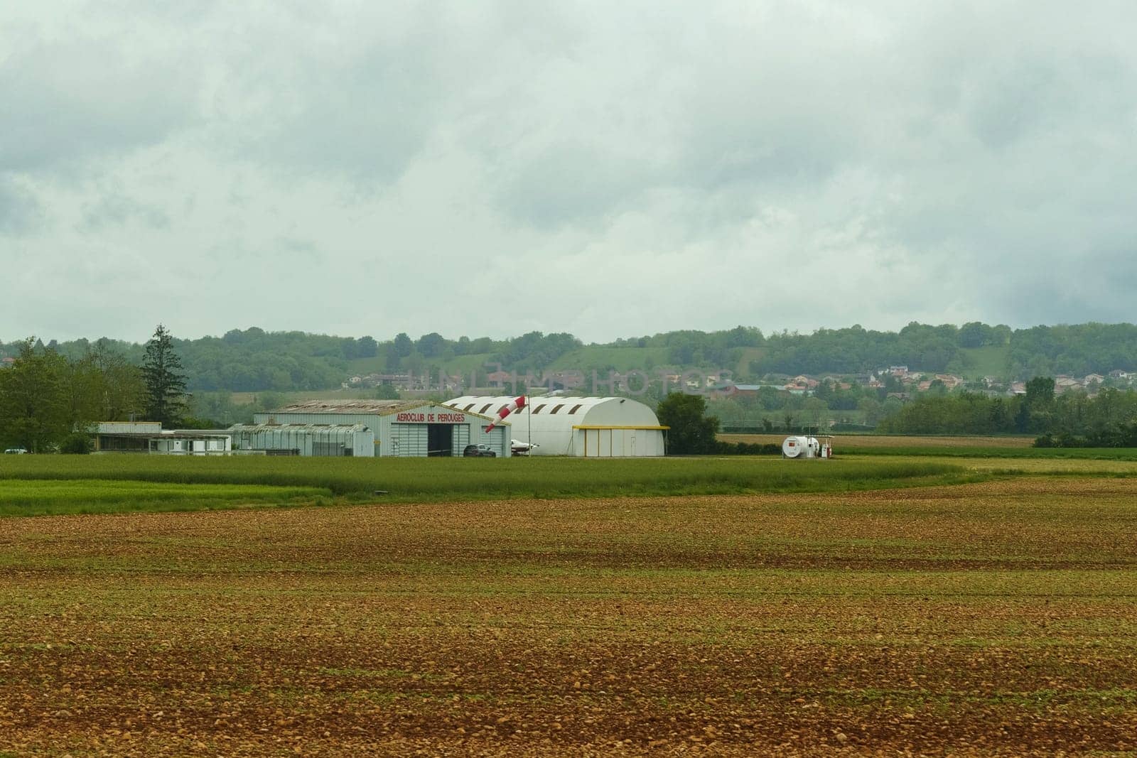 Lyon, France - May 7, 2023: Aeroclub de Perouges hangars in a rural French landscape, surrounded by fields and greenery, under a cloudy spring sky.