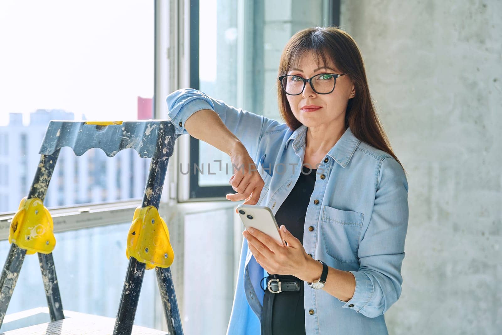 Female decorator designer, repair manager, organizer of final finishing and decor with smartphone, looking at camera in new apartment office. Repair, decoration, work, people concept