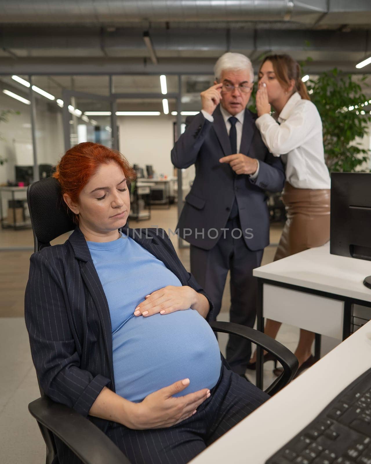 Elderly Caucasian man and woman gossiping behind their sleeping pregnant colleague in the office. Vertical photo