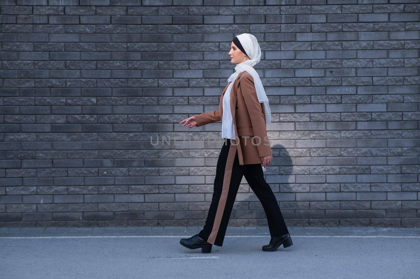 A young woman dressed in a hijab and a business suit walks along a brick wall. by mrwed54