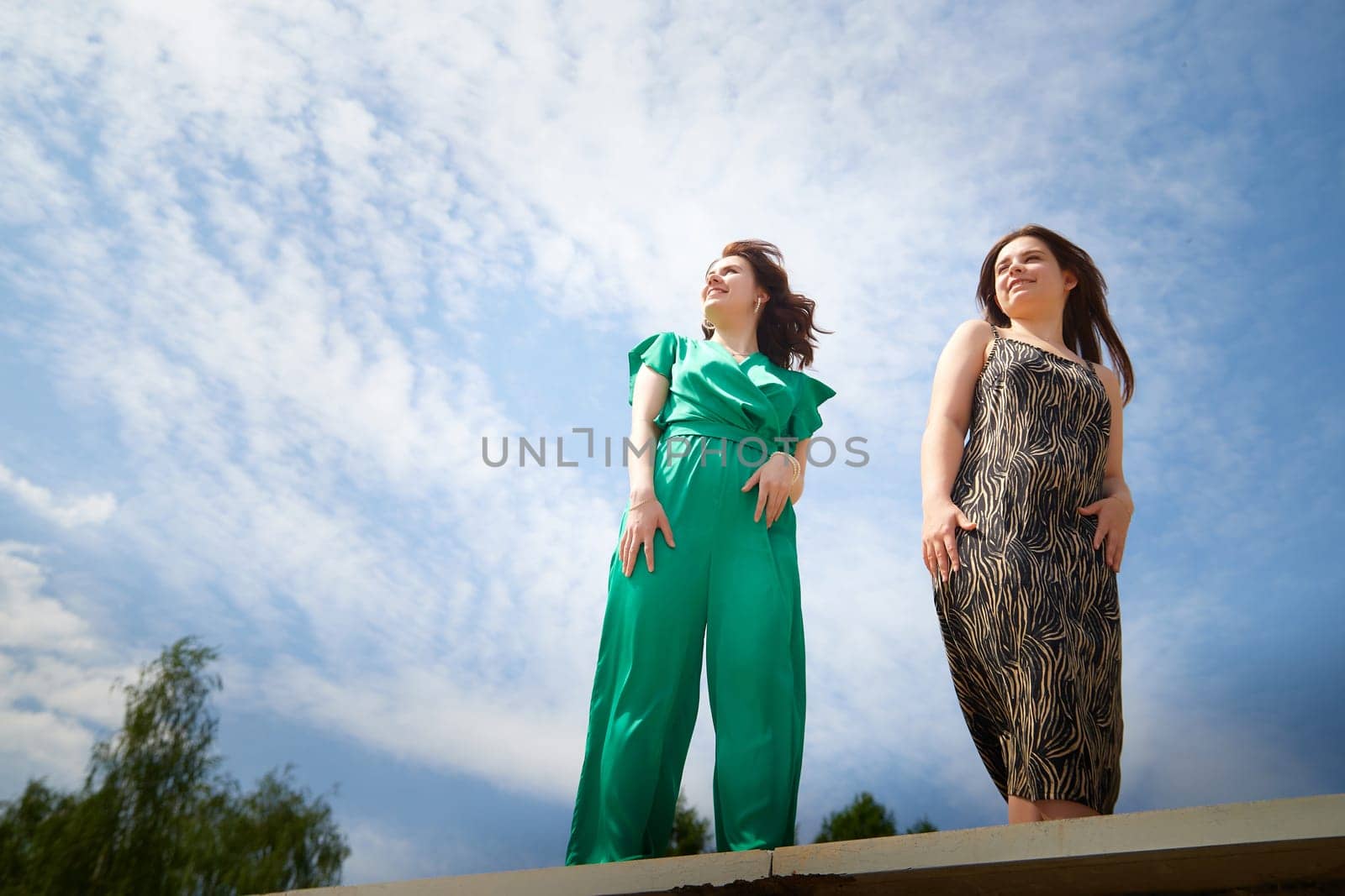 Happy Girls Enjoying Summer Day Together in the Park. Two young women stand on a platform in a park under a bright summer sky, smiling and relaxed