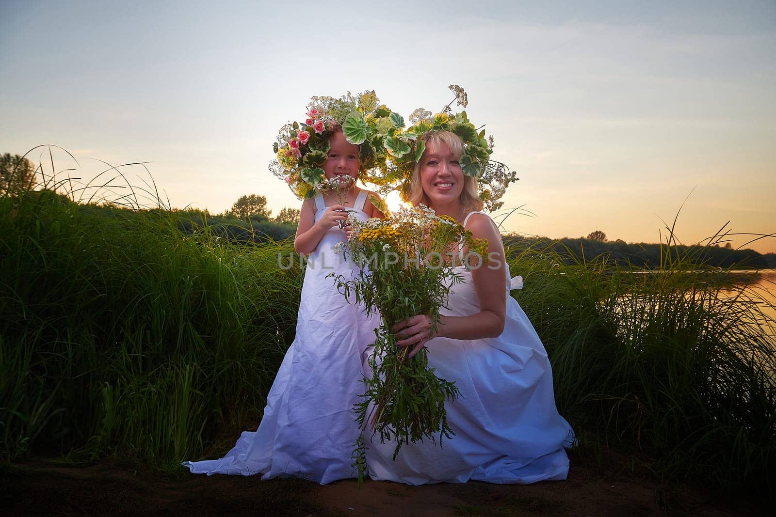 Mother and daughter in white sundresses, wreaths of flowers and bouquet on nature in evening at sunset. Family celebrate Slavic Holiday of Ivan Kupala by keleny