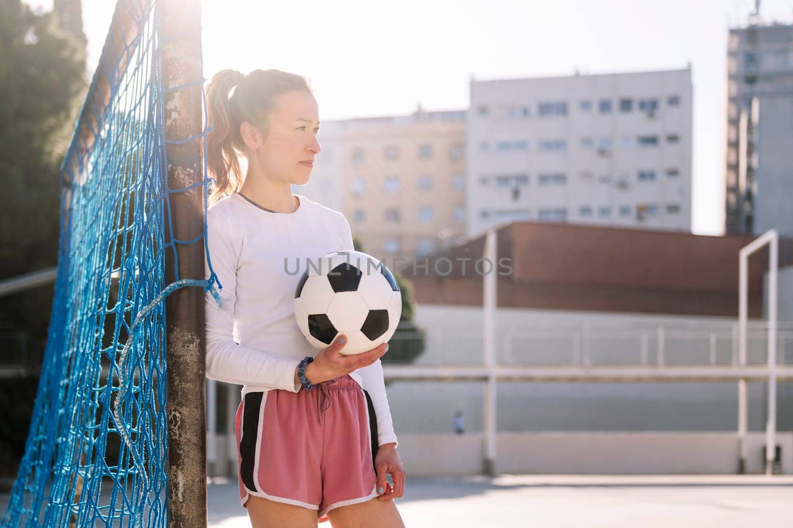 caucasian female football player holding a soccer ball leaning on the goal of an urban court, concept of sport and active lifestyle, copy space for text