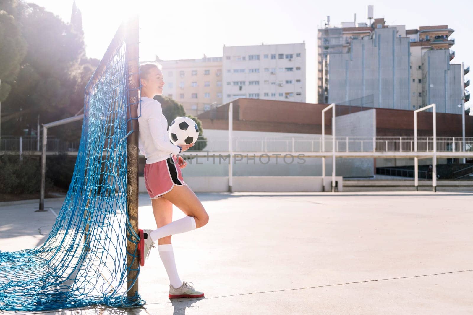smiling caucasian female football player holding a soccer ball leaning on the goal of an urban court, concept of sport and active lifestyle, copy space for text