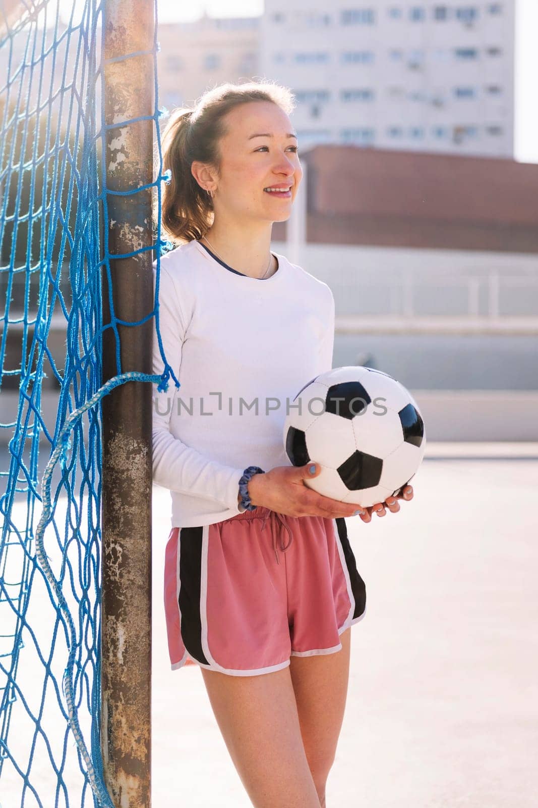 caucasian female football player holding a soccer ball leaning on the goal of an urban court, concept of sport and active lifestyle